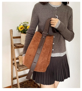Brown suede bucket bag with wide leather strap held by a model in a minimalist setting