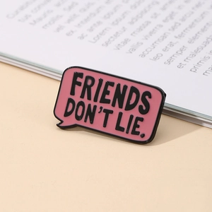 Pink enamel speech bubble pin with black Friends Don't Lie text on beige book surface