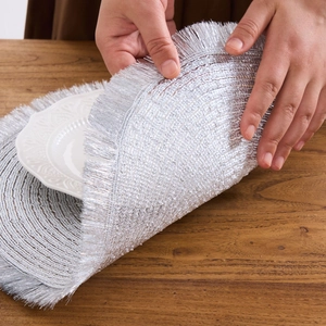 Detail of silver round placemat with fringe texture held by hands on wooden table