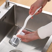 Long-handled white cleaning brush scrubbing the inside of a clear glass in a kitchen sink