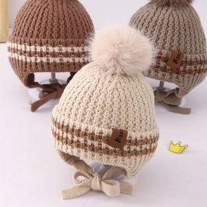 Close-up of knit baby earflap hats in brown and beige with pom-poms and Kuki labels
