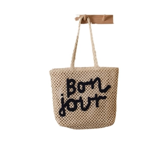 Straw woven tote bag with Bonjour embroidery hanging on a wooden hook for beach vacation