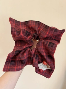 Detail of red and black checkered scrunchie with HAPPY label held in hand on neutral backdrop