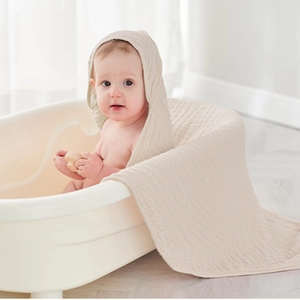 Close-up of a baby in a white bathtub wearing a soft beige 6-layer cotton muslin hooded towel