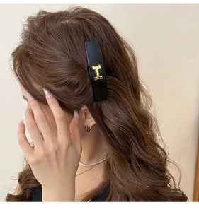 Close-up of black acrylic hair clip with a gold H-shaped metal letter on brown curly hair