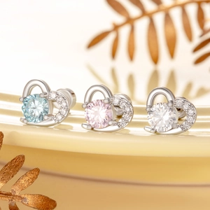 Three heart-shaped stud earrings with blue, pink, and white zircons on a beige reflective surface