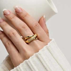 Close-up of a hand holding a white cup, wearing a gold X-shaped ring with pavé zircons