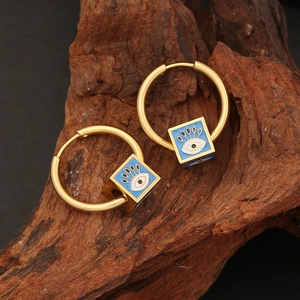 Golden hoop earrings with blue Evil Eye square charms on a textured wood background