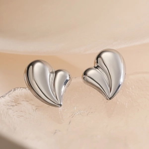 Minimalist silver heart stud earrings with a polished 3D finish on a warm textured background