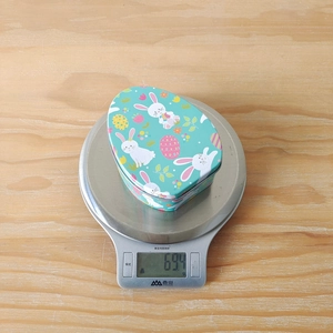 Flat egg-shaped Easter tin box with rabbit print shown on a digital scale weighing 69.4g