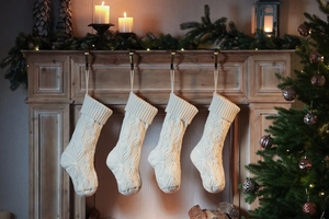 Four white knitted Christmas stockings with cable patterns hanging on a decorated fireplace