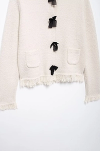 Detail of cream knit cardigan featuring black bow buttons and elegant tassel trim accents