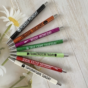 Set of 7 glitter ballpoint pens with weekday themes on a light wood background
