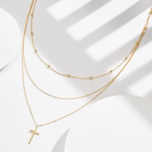 Three-layer gold beaded chain necklace with cross pendant on minimalist background