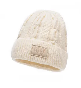 Dettaglio del berretto in maglia beige con trama a trecce spesse e patch logo in pelle frontale