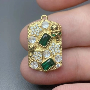 Gold rectangular pendant with green emerald gems and skull detail held by hand