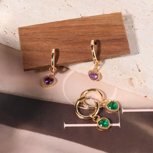 Elegant gold heart-shaped earrings with purple and green stones on a wooden block