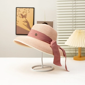 Detail of a beige woven straw hat featuring a pink ribbon and a gold 'M' metal badge