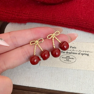 Detail view of red cherry earrings with gold bow accents held by a hand on a knit background