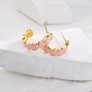 Gold C-shaped hoop earrings with pink enamel smileys on a white stone background