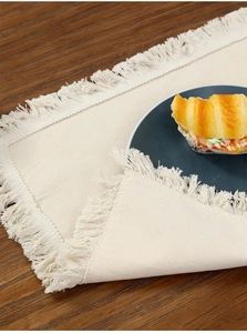 Detail of cream cotton linen placemat with tassel edges on dark wooden dining table