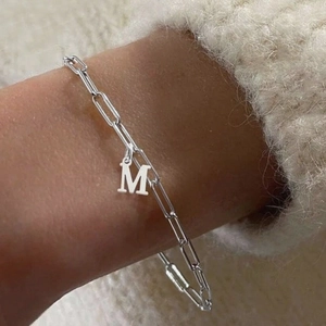 Silver paperclip link bracelet with an 'M' initial pendant worn on wrist over a beige sweater