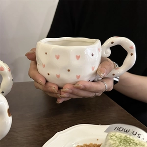 Detail of irregular ceramic coffee mug with pink hearts and wavy handle held by hands