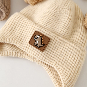 Beige baby knit hat with double pom poms and cute dinosaur patch for winter warmth