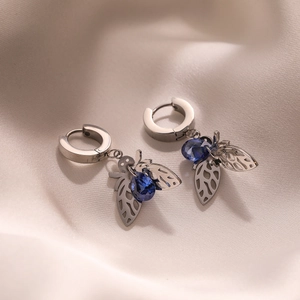 Silver fly earrings with blue crystal wing charms on soft beige silk background