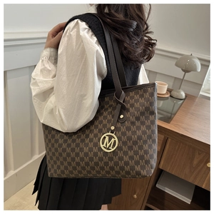 Detail of a brown tote bag with geometric M-pattern and gold charm on a shoulder