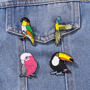 Set of four colorful bird enamel pins featuring parrots and toucans on denim jacket